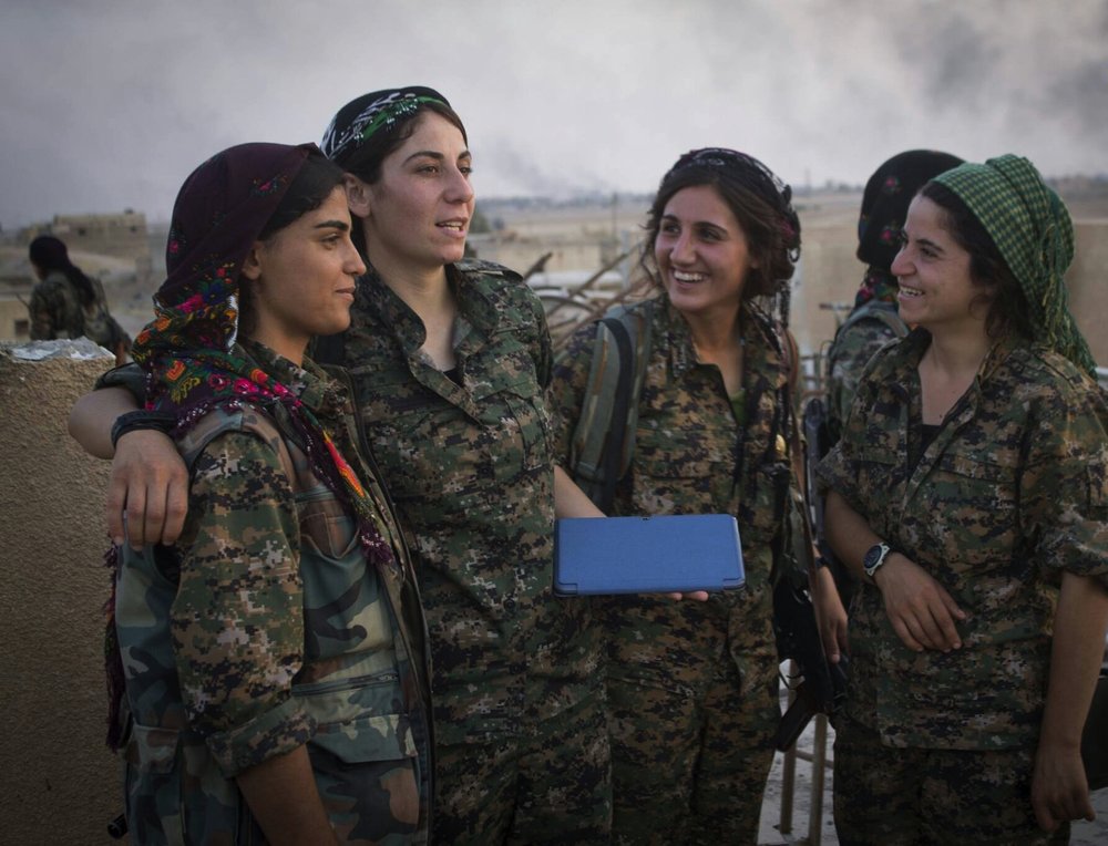 Four YPJ women in camouflage uniforms, one holding a tablet, smoke on the horizon