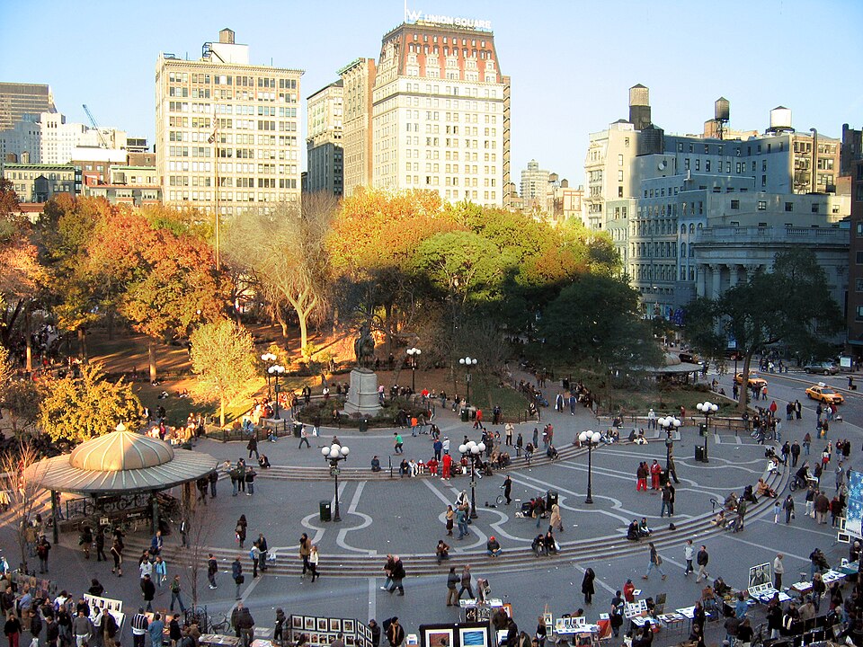 Union Square, New York City, in autumn. An elevated view of the south plaza shows people moving across the wave-patterned paving, vendors displaying artwork along the edges, the Lincoln statue at center, and the W Union Square building rising behind.